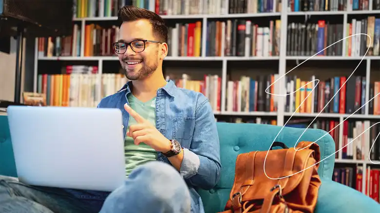 Estudiante masculino sonriente en la biblioteca con un libro en la mano y una computadora en frente tomando una clase en Ciencias de la Educación.