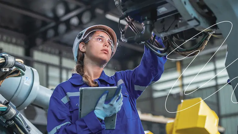 Una mujer con casco y gafas de seguridad sosteniendo un papel mira atentamente hacia una máquina en fábrica.